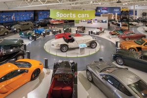 Interior view of British Motor Museum. A variety of classic and antique sports cars circle the room, with a white an red car in the centre.